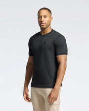 A man wearing the Psycho Bunny MENS BRYCE PERFORMANCE TEE (B6U490F200) in black and beige pants stands against a plain white background, exuding effortless style and comfort.