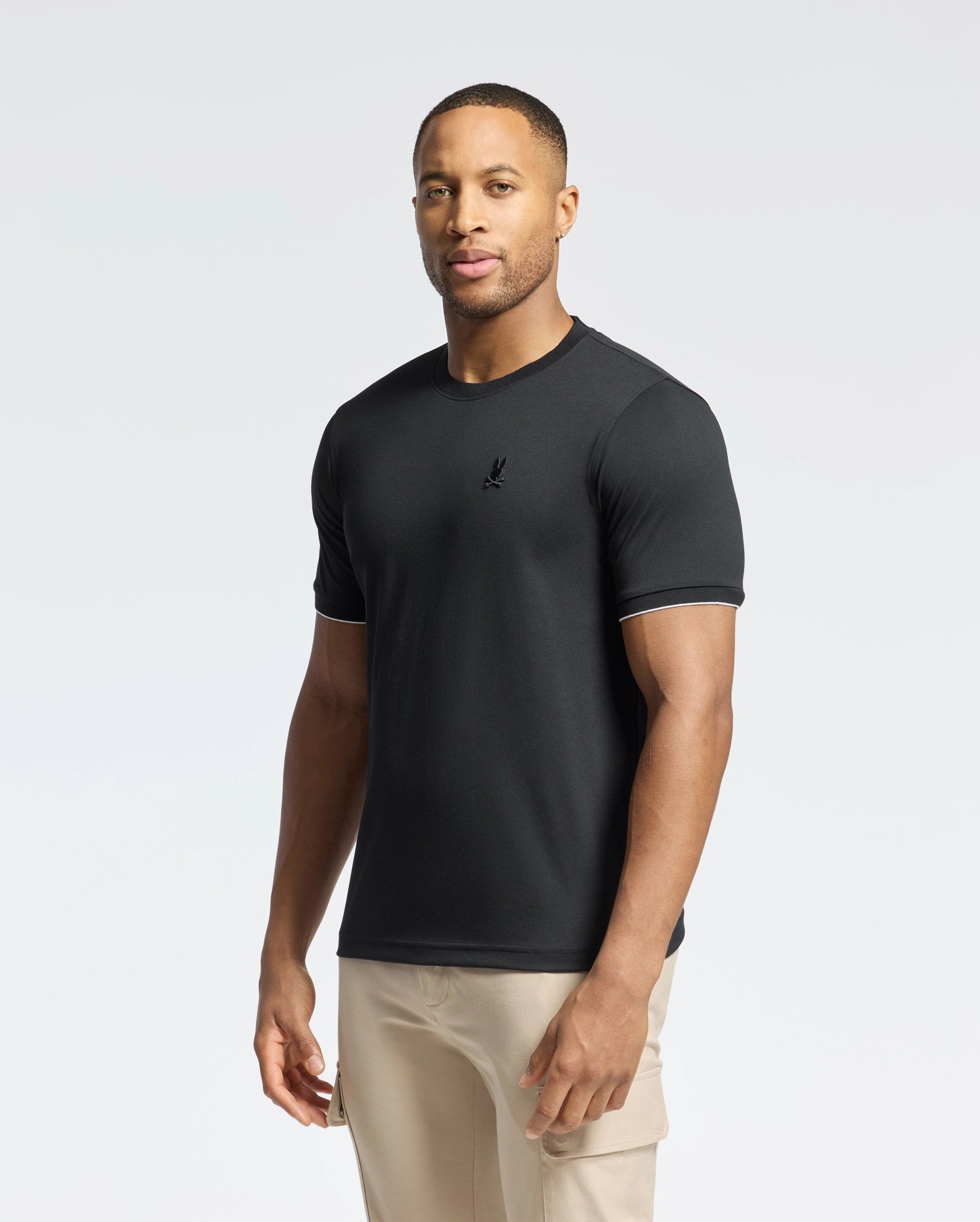 A man wearing the Psycho Bunny MENS BRYCE PERFORMANCE TEE (B6U490F200) in black and beige pants stands against a plain white background, exuding effortless style and comfort.