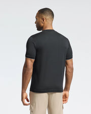 A man faces away wearing a Psycho Bunny MENS BRYCE PERFORMANCE TEE in black with white trim, made from pique fabric, paired with beige cargo shorts. The background is light gray.