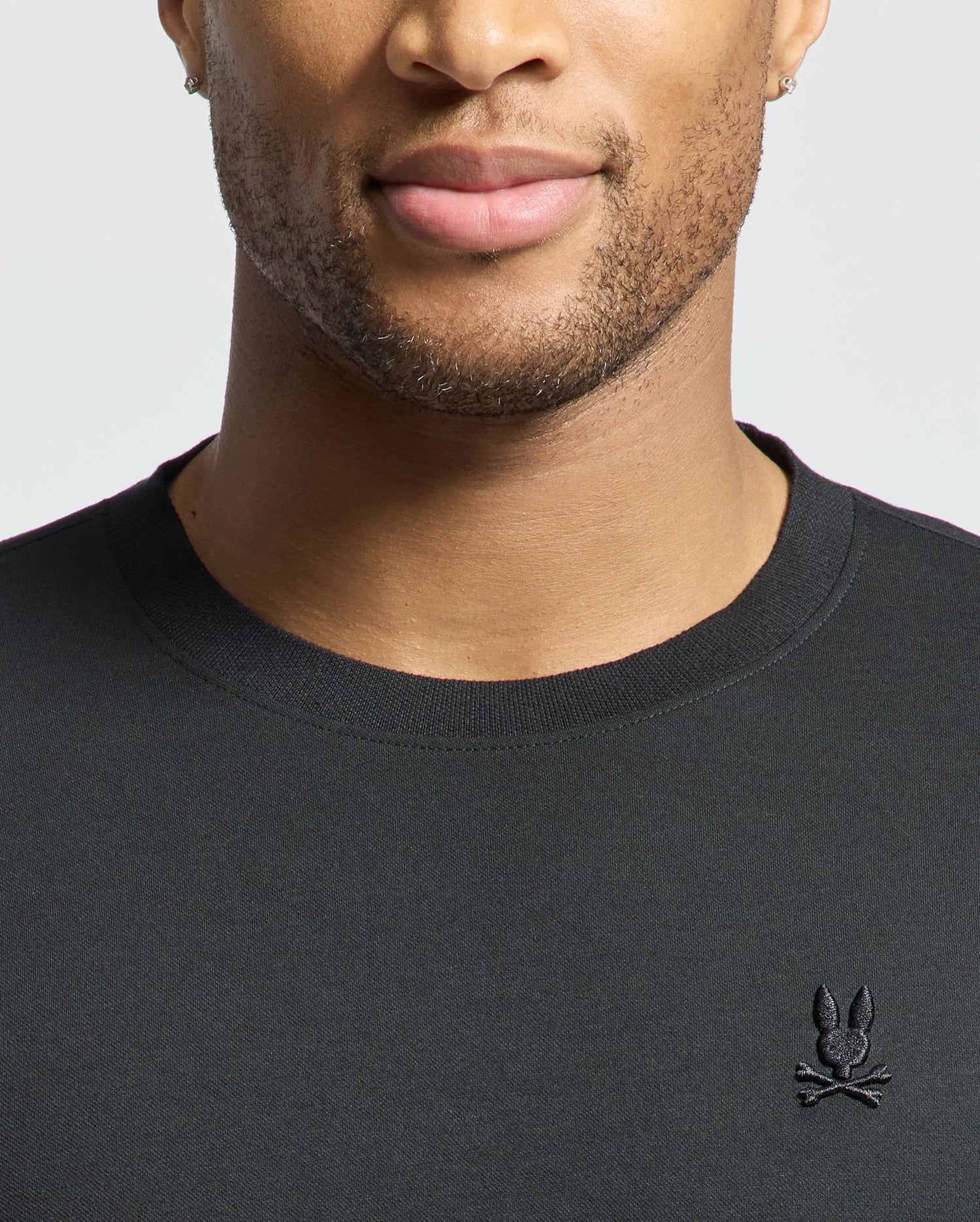 A close-up shows a man in a Psycho Bunny MENS BRYCE PERFORMANCE TEE (B6U490F200), made from Modal-Pima Cotton. The black tee features an embroidered bunny and crossbones logo on the chest, highlighting the luxurious Peruvian fabric from his lower face to shoulders.