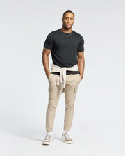 A man in a black Psycho Bunny MENS BRYCE PERFORMANCE TEE (B6U490F200), beige cargo pants, and beige sneakers stands on a plain white background. He has a light gray sweater tied around his waist, with hands in pockets, confidently looking at the camera.