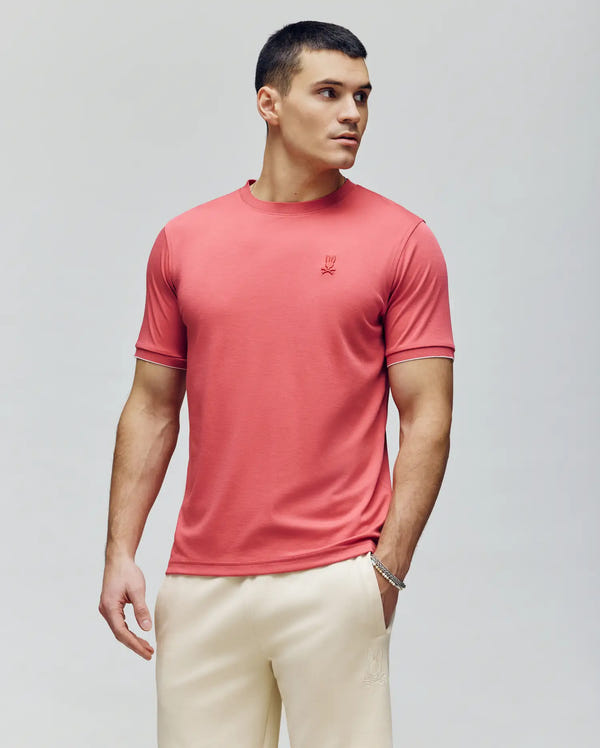 A man with short dark hair wears the Psycho Bunny MENS BRYCE PERFORMANCE TEE - B6U490F200 in coral and cream pants, standing against a plain light background, gazing to the side with a neutral expression and one hand in his pocket.
