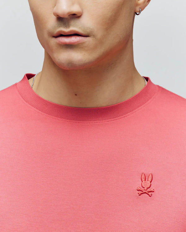 A person wearing a pink Psycho Bunny MENS BRYCE PERFORMANCE TEE (B6U490F200) with an embroidered bunny and crossbones logo; only the lower half of their face and upper torso are visible.