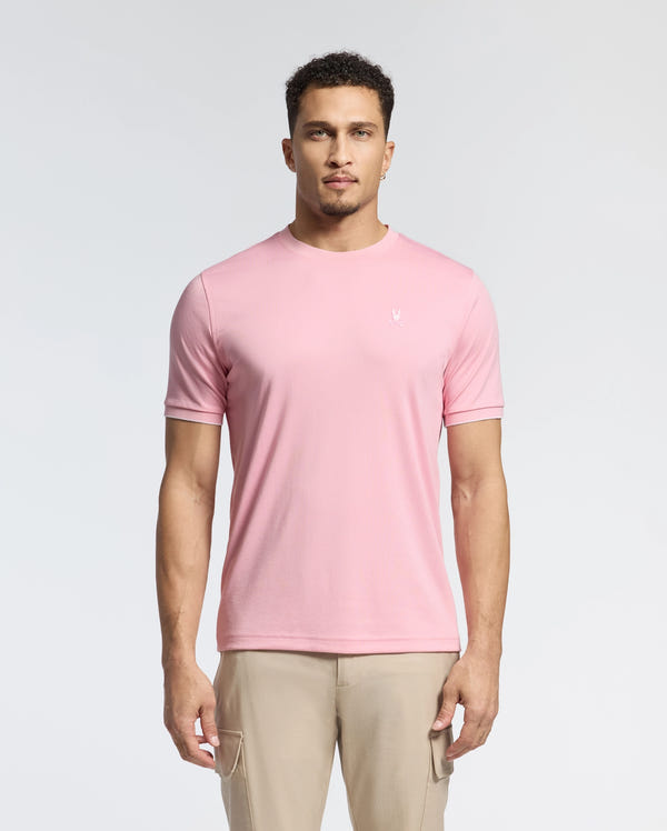 A man in a light pink Psycho Bunny Men's Bryce Performance Tee (B6U490F200), made from a Modal-Pima cotton blend in Peru, stands against a white background. He has short curly hair and a neutral expression. The tee features a small embroidered logo on the left chest.