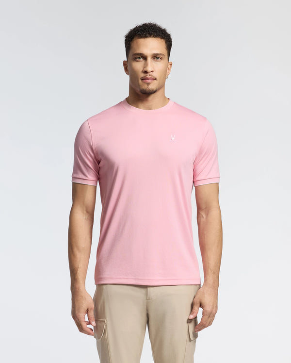 A man in a light pink Psycho Bunny Men's Bryce Performance Tee (B6U490F200), made from a Modal-Pima cotton blend in Peru, stands against a white background. He has short curly hair and a neutral expression. The tee features a small embroidered logo on the left chest.