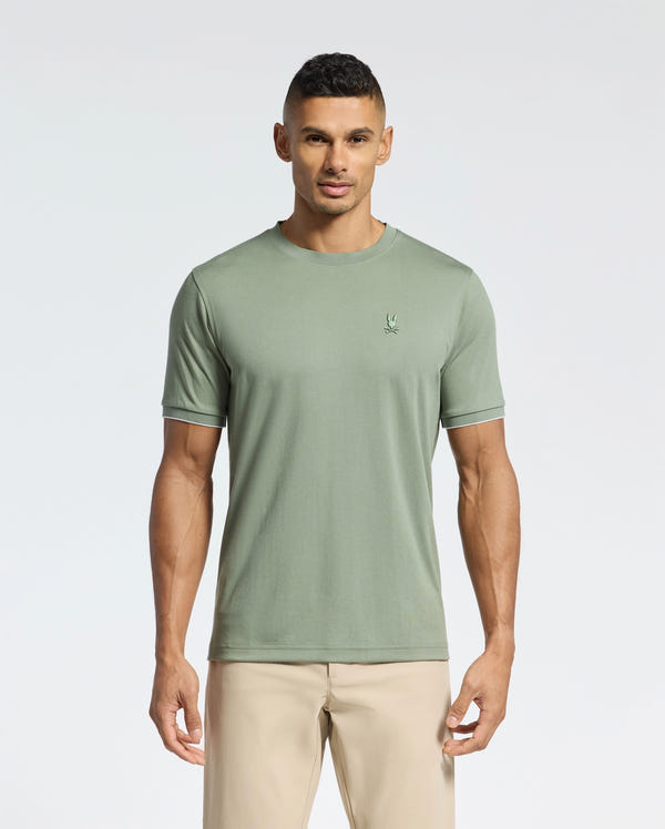 A man wears a light green MENS BRYCE PERFORMANCE TEE by Psycho Bunny and beige pants against a white background. Made from soft Pima cotton with Peruvian craftsmanship, the tee has a small embroidered chest logo. He looks forward neutrally.
