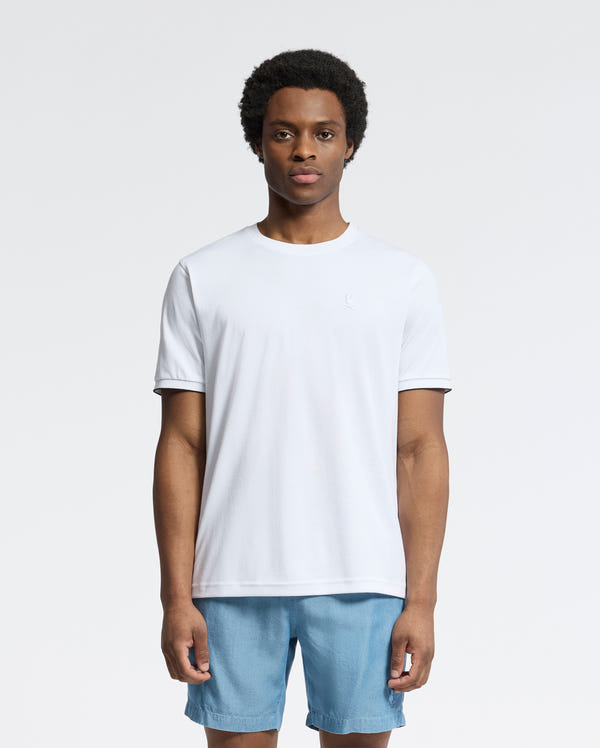 A person stands against a white background wearing the Psycho Bunny MENS BRYCE PERFORMANCE TEE - B6U490F200, a slim fit plain white T-shirt made from a Modal-Pima cotton blend, paired with light blue shorts.