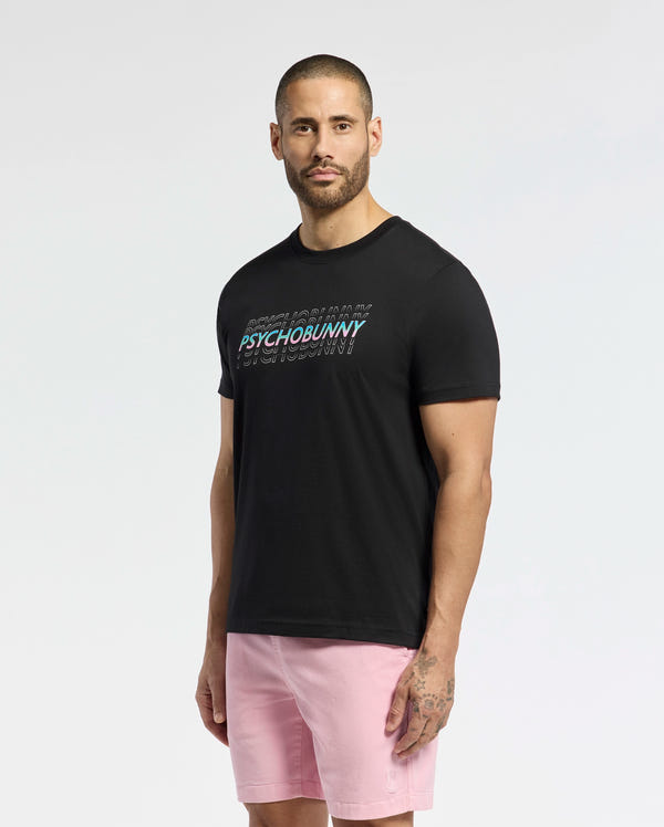 A man with short hair and a beard wears the Psycho Bunny MENS CANBERRA GRAPHIC TEE (B6U526F200) in black and pink shorts, standing against a plain white background.