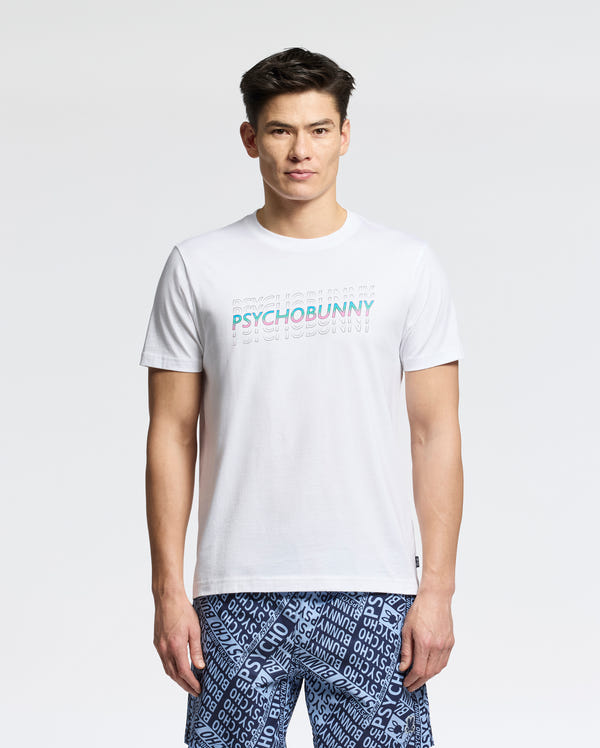A man stands facing forward wearing the Psycho Bunny MENS CANBERRA GRAPHIC TEE (B6U526F200), a white t-shirt with colorful "PSYCHOBUNNY" lettering, paired with blue shorts featuring a white text pattern. The background is plain and light-colored.