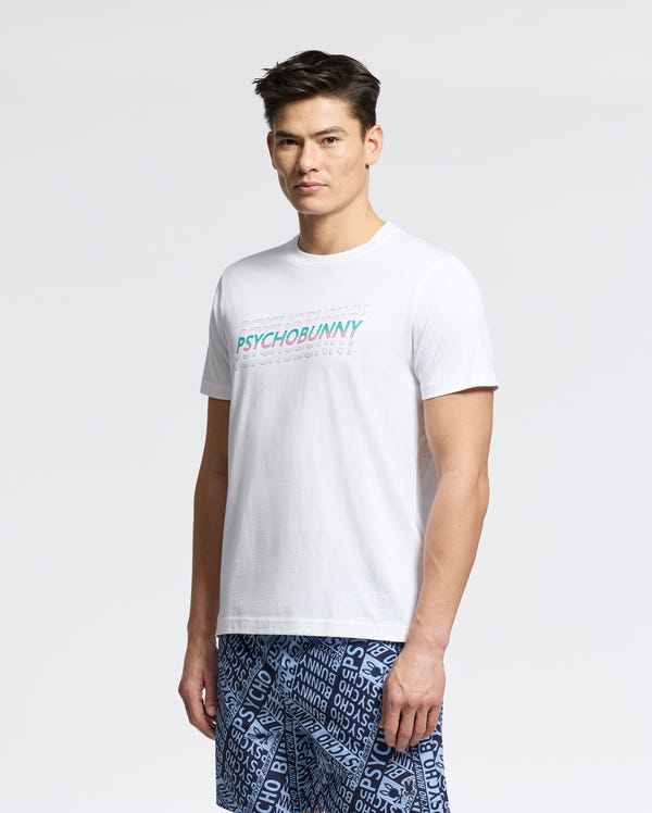 A man with short dark hair wears the MENS CANBERRA GRAPHIC TEE (B6U526F200) by Psycho Bunny, featuring colorful text, paired with blue patterned shorts, standing against a plain light gray background.