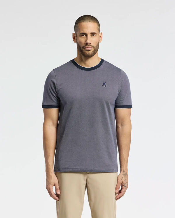 A man with a beard stands facing forward in a Psycho Bunny MENS MACKAY JACQUARD FASHION TEE (B6U576F200), featuring dark trim and an embroidered chest Bunny logo, paired with light-colored pants against a plain, light background.