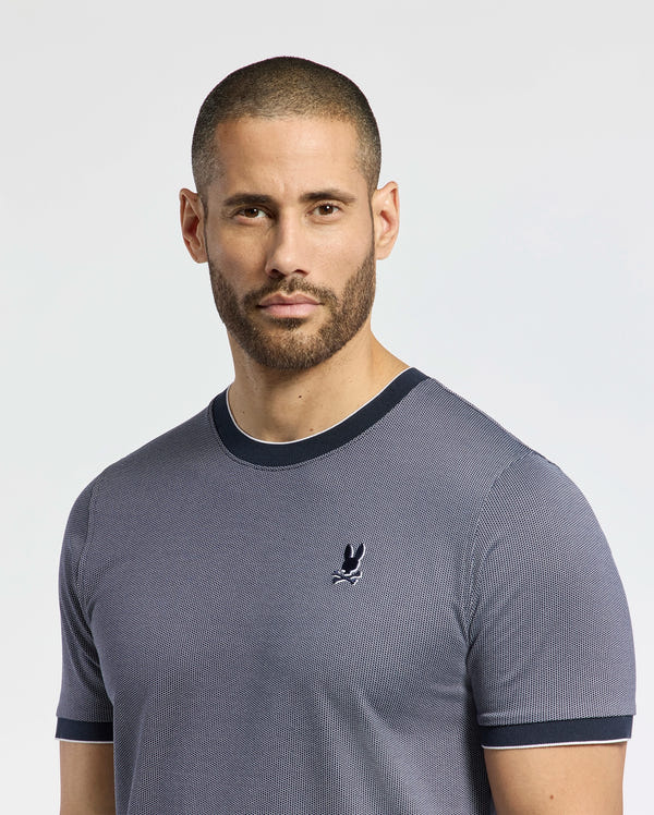 A short-haired, bearded individual dons a Psycho Bunny MENS MACKAY JACQUARD FASHION TEE (B6U576F200) in gray, ribbed Jacquard-knit Pima cotton with an embroidered chest Bunny logo, set against a plain light background that enhances its premium quality.