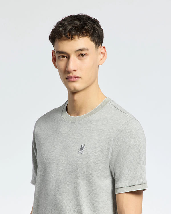 A person with short dark hair is wearing the light gray MENS MACKAY JACQUARD FASHION TEE by Psycho Bunny. It features a premium Pima cotton material and an embroidered chest Bunny. The background is plain white, and they are looking directly at the camera.