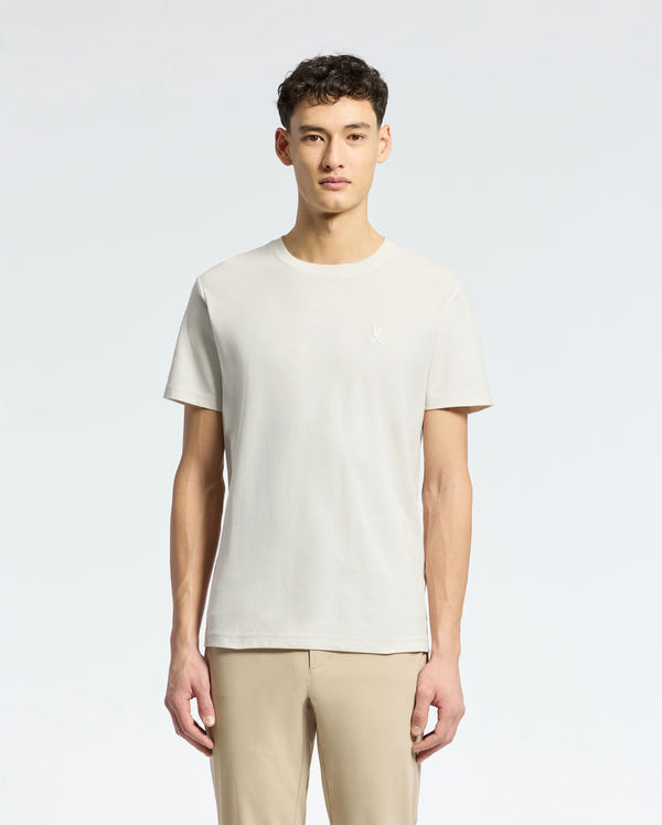 Amidst a light grey background, an individual with short curly dark hair dons a Psycho Bunny MENS TAYLOR PIQUE TEE in light beige. The regularly fit tee is made from lightweight Pima cotton and paired with beige pants, while facing forward.