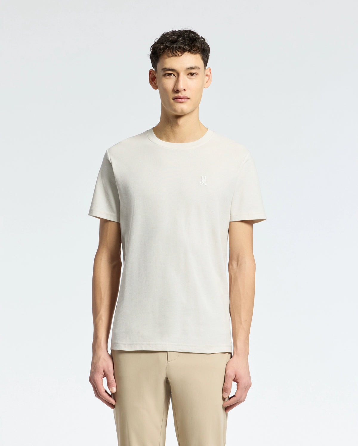 Amidst a light grey background, an individual with short curly dark hair dons a Psycho Bunny MENS TAYLOR PIQUE TEE in light beige. The regularly fit tee is made from lightweight Pima cotton and paired with beige pants, while facing forward.