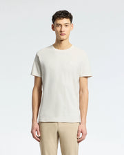 Amidst a light grey background, an individual with short curly dark hair dons a Psycho Bunny MENS TAYLOR PIQUE TEE in light beige. The regularly fit tee is made from lightweight Pima cotton and paired with beige pants, while facing forward.