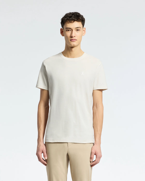 Amidst a light grey background, an individual with short curly dark hair dons a Psycho Bunny MENS TAYLOR PIQUE TEE in light beige. The regularly fit tee is made from lightweight Pima cotton and paired with beige pants, while facing forward.
