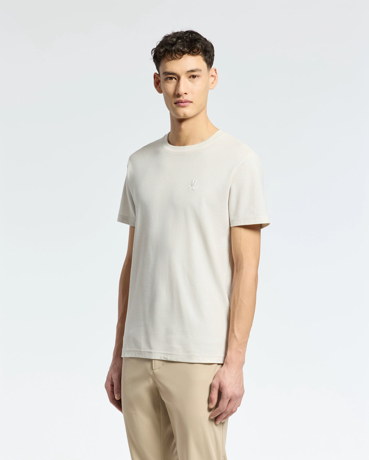 A person stands against a light background wearing the Psycho Bunny MENS TAYLOR PIQUE TEE - B6U579F200, a plain beige lightweight Pima cotton t-shirt with an embroidered chest Bunny logo on the left side, paired with tan pants. Their arms are relaxed at their sides.