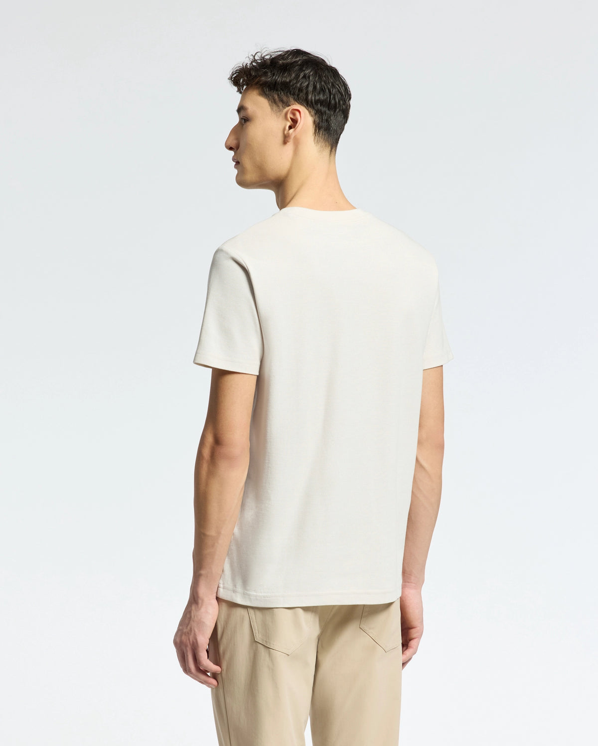 A person stands with their back to the camera, wearing a light beige Psycho Bunny MENS TAYLOR PIQUE TEE, crafted from lightweight Pima cotton, and tan pants. The regular fit tee features a subtle embroidered chest Bunny. The background is a plain, light color.