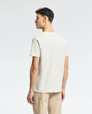 A person stands with their back to the camera, wearing a light beige Psycho Bunny MENS TAYLOR PIQUE TEE, crafted from lightweight Pima cotton, and tan pants. The regular fit tee features a subtle embroidered chest Bunny. The background is a plain, light color.