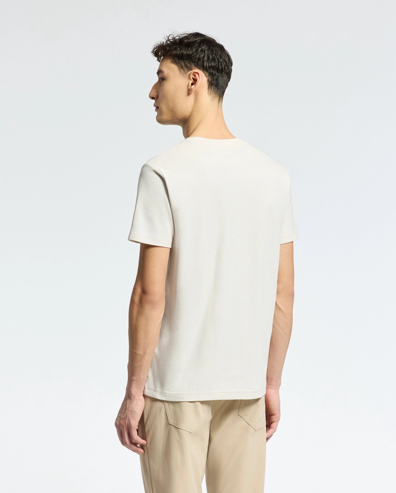 A person stands with their back to the camera, wearing a light beige Psycho Bunny MENS TAYLOR PIQUE TEE, crafted from lightweight Pima cotton, and tan pants. The regular fit tee features a subtle embroidered chest Bunny. The background is a plain, light color.