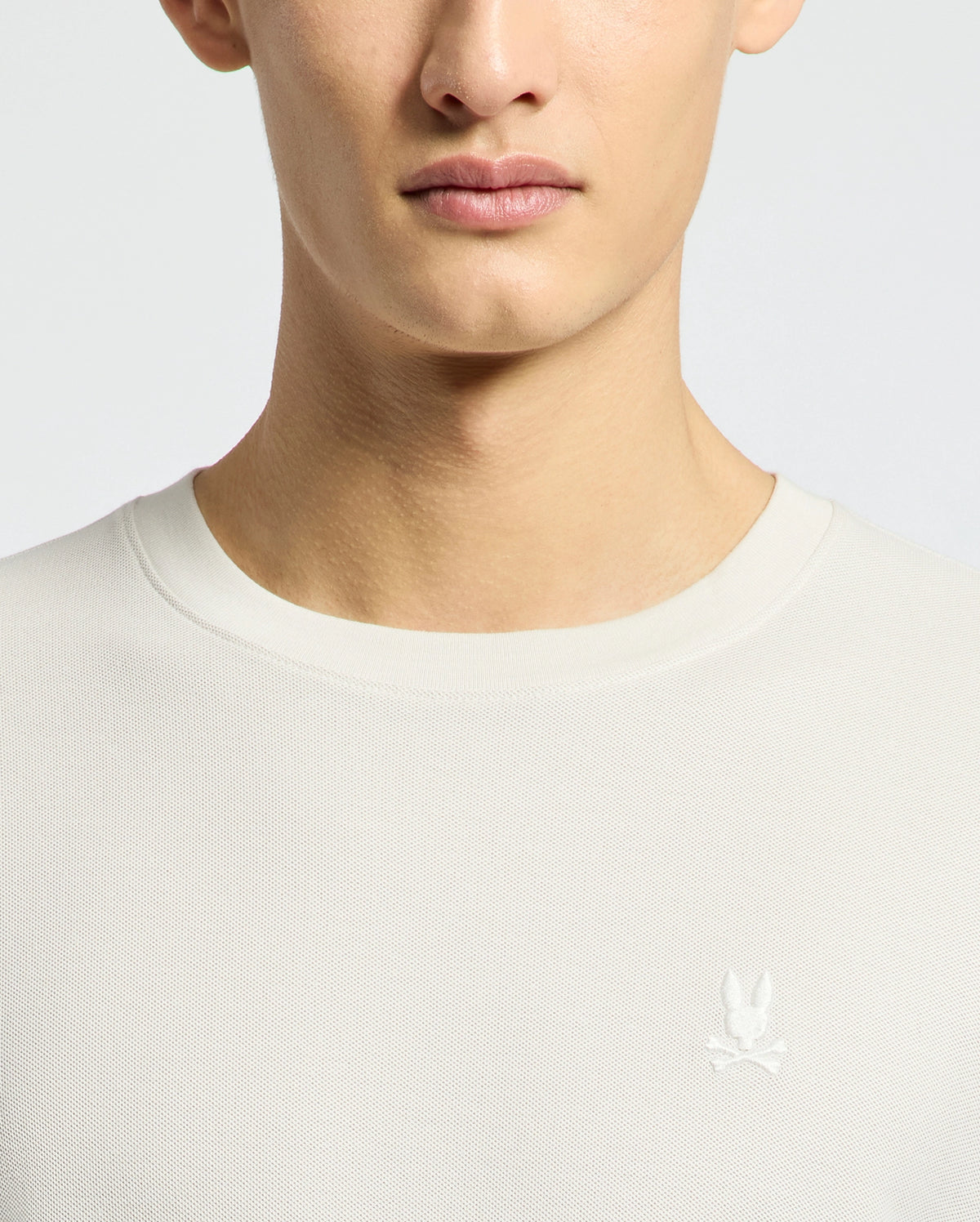 A person models the MENS TAYLOR PIQUE TEE - B6U579F200 by Psycho Bunny, wearing a regular fit white crew-neck shirt featuring an embroidered chest bunny logo. The lightweight Pima cotton is highlighted, with focus on the upper torso and part of the face against a light backdrop.