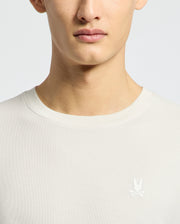 A person models the MENS TAYLOR PIQUE TEE - B6U579F200 by Psycho Bunny, wearing a regular fit white crew-neck shirt featuring an embroidered chest bunny logo. The lightweight Pima cotton is highlighted, with focus on the upper torso and part of the face against a light backdrop.