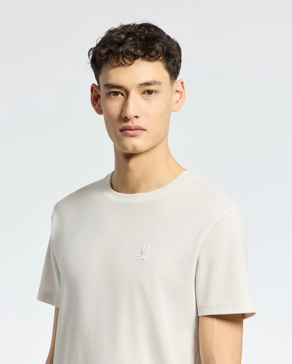 A person with short, dark curly hair is wearing the light gray MENS TAYLOR PIQUE TEE by Psycho Bunny, a regular fit made from lightweight Pima cotton featuring an embroidered chest Bunny logo, standing against a plain light background.