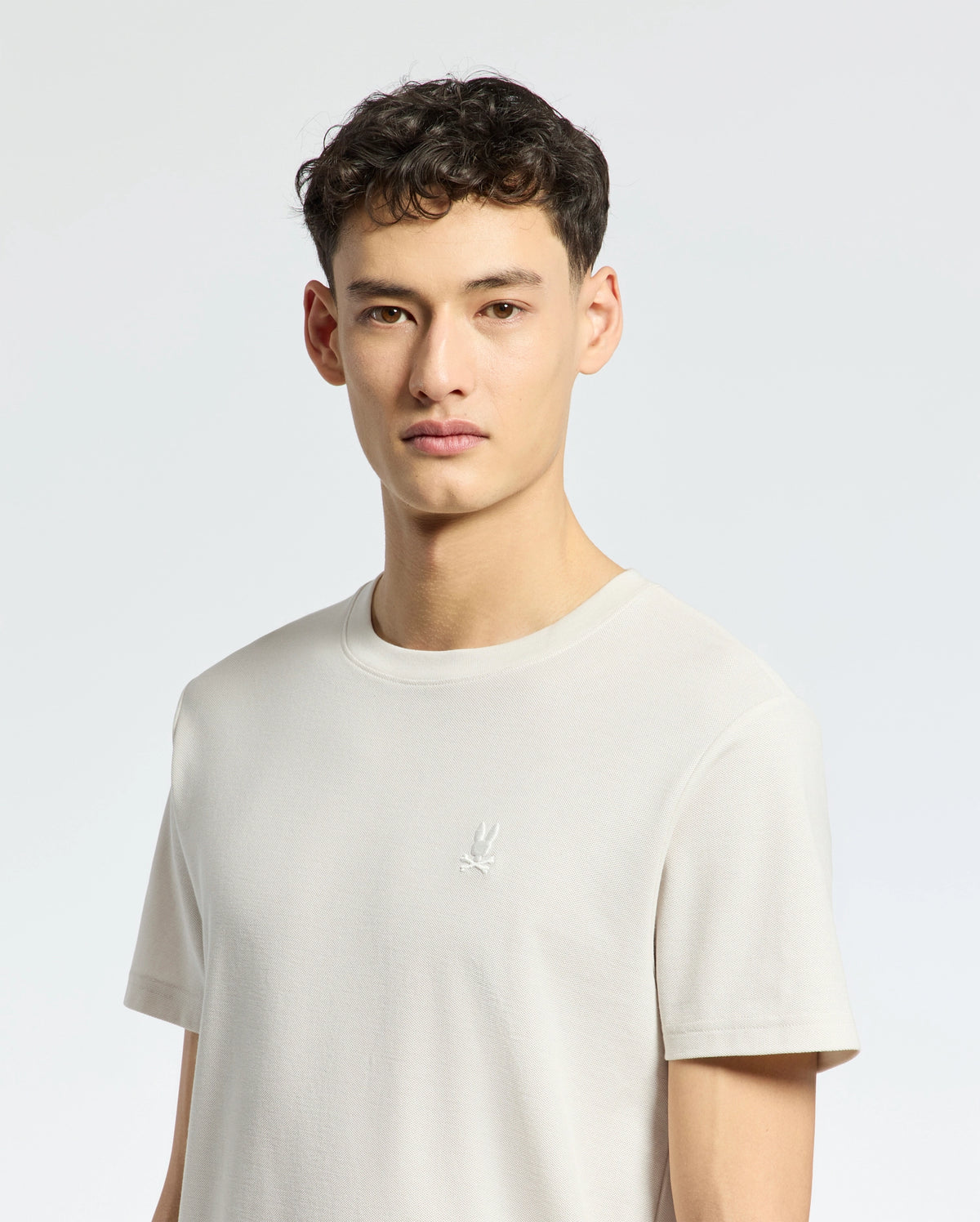 A person with short, dark curly hair is wearing the light gray MENS TAYLOR PIQUE TEE by Psycho Bunny, a regular fit made from lightweight Pima cotton featuring an embroidered chest Bunny logo, standing against a plain light background.