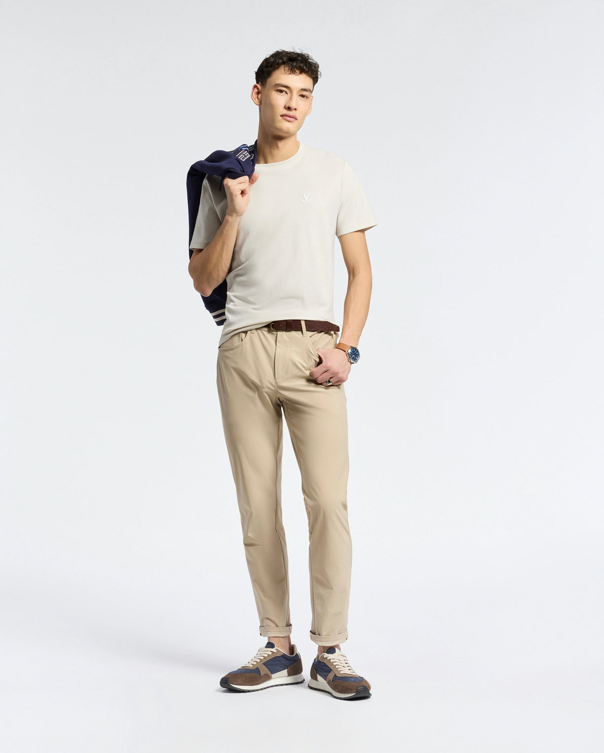 A man in a Psycho Bunny MENS TAYLOR PIQUE TEE (B6U579F200), light beige with an embroidered chest Bunny logo, and tan pants holds a dark jacket over his shoulder. He sports brown/blue sneakers and a watch, standing against a white backdrop. The regular-fit tee is made from lightweight Pima cotton for comfort.