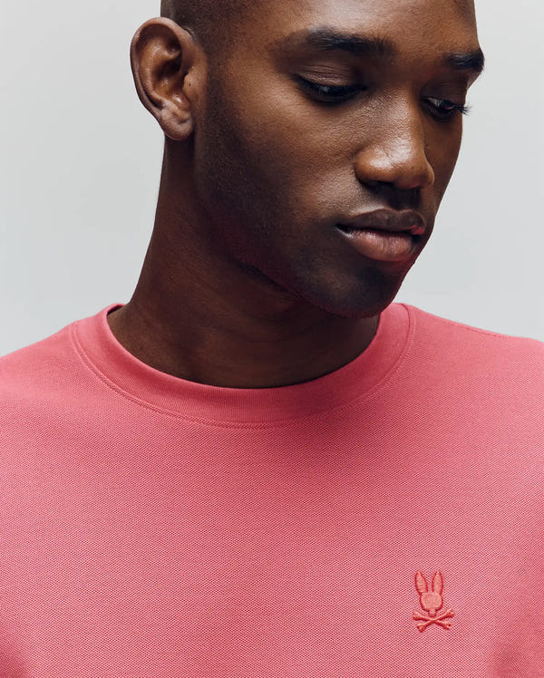 A man in a pink Psycho Bunny MENS TAYLOR PIQUE TEE (B6U579F200) with a small embroidered bunny and crossbones logo looks down neutrally against a light gray background.