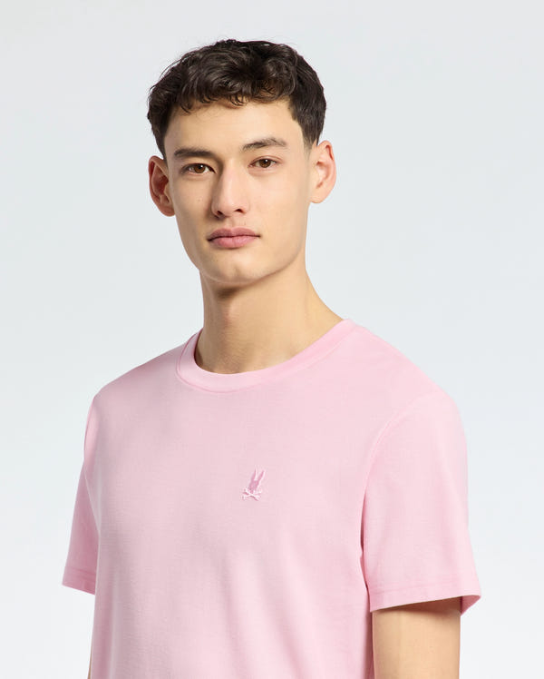 A person in a Psycho Bunny MENS TAYLOR PIQUE TEE - B6U579F200, featuring an embroidered chest Bunny, stands against a light gray backdrop. The individual with short dark hair looks directly at the camera.