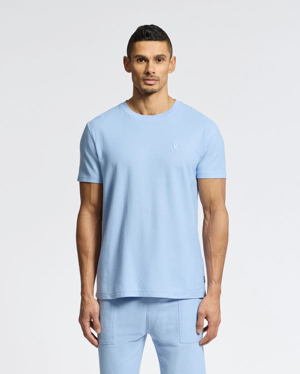 A man in a light blue Psycho Bunny Men's Taylor Pique Tee - B6U579F200 and matching pants stands against a white backdrop. The lightweight piqué tee features an embroidered Bunny on the left chest, complementing his short dark hair and goatee.