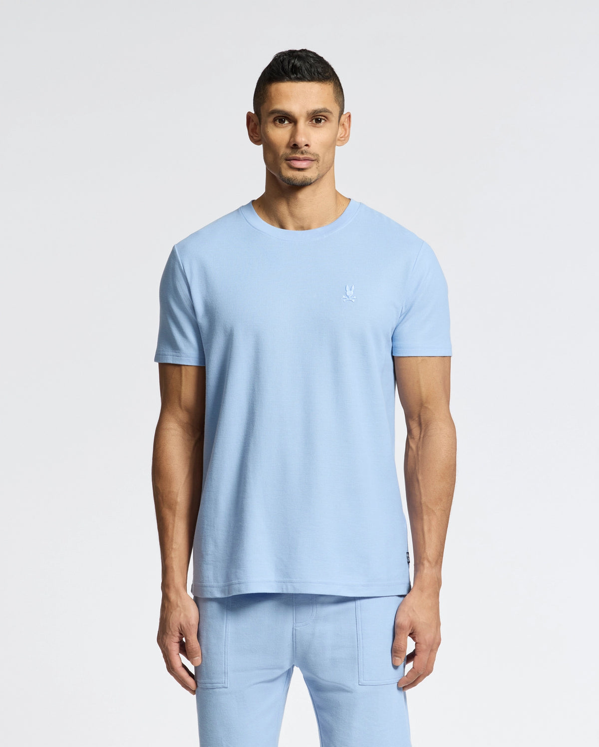A man in a light blue Psycho Bunny Men's Taylor Pique Tee - B6U579F200 and matching pants stands against a white backdrop. The lightweight piqué tee features an embroidered Bunny on the left chest, complementing his short dark hair and goatee.