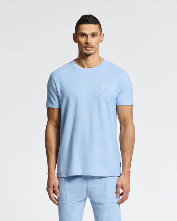 A man in a light blue Psycho Bunny Men's Taylor Pique Tee - B6U579F200 and matching pants stands against a white backdrop. The lightweight piqué tee features an embroidered Bunny on the left chest, complementing his short dark hair and goatee.