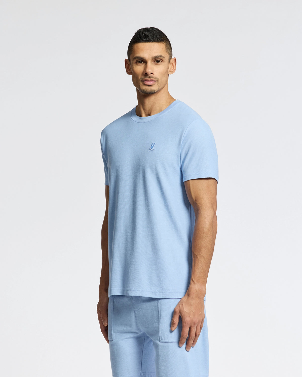 A man in a light blue Psycho Bunny MENS TAYLOR PIQUE TEE - B6U579F200 and matching shorts stands against a plain white background. With short, dark hair and a neutral expression, the lightweight piqué fabric subtly highlights his style.