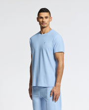 A man in a light blue Psycho Bunny MENS TAYLOR PIQUE TEE - B6U579F200 and matching shorts stands against a plain white background. With short, dark hair and a neutral expression, the lightweight piqué fabric subtly highlights his style.