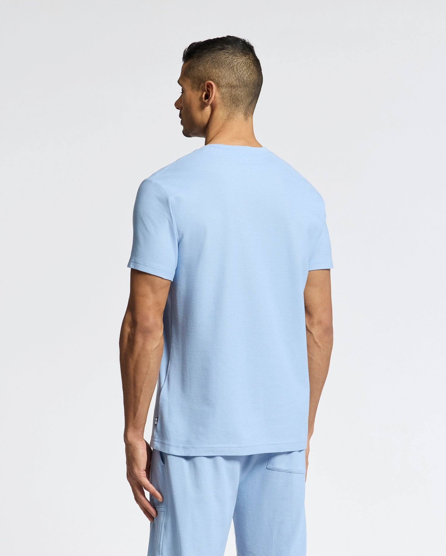 A man with short dark hair faces away from the camera, wearing a Psycho Bunny MENS TAYLOR PIQUE TEE - B6U579F200 in light blue and matching shorts against a plain white background.