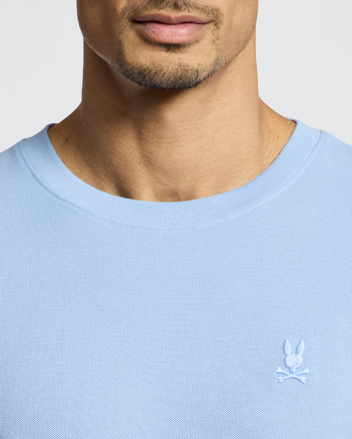 A person wears the Psycho Bunny MENS TAYLOR PIQUE TEE, a light blue Pima cotton shirt with textured fabric and a round neckline, showcasing a small white embroidered bunny logo on the chest. The image highlights the chest and neck area.
