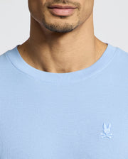 A person wears the Psycho Bunny MENS TAYLOR PIQUE TEE, a light blue Pima cotton shirt with textured fabric and a round neckline, showcasing a small white embroidered bunny logo on the chest. The image highlights the chest and neck area.