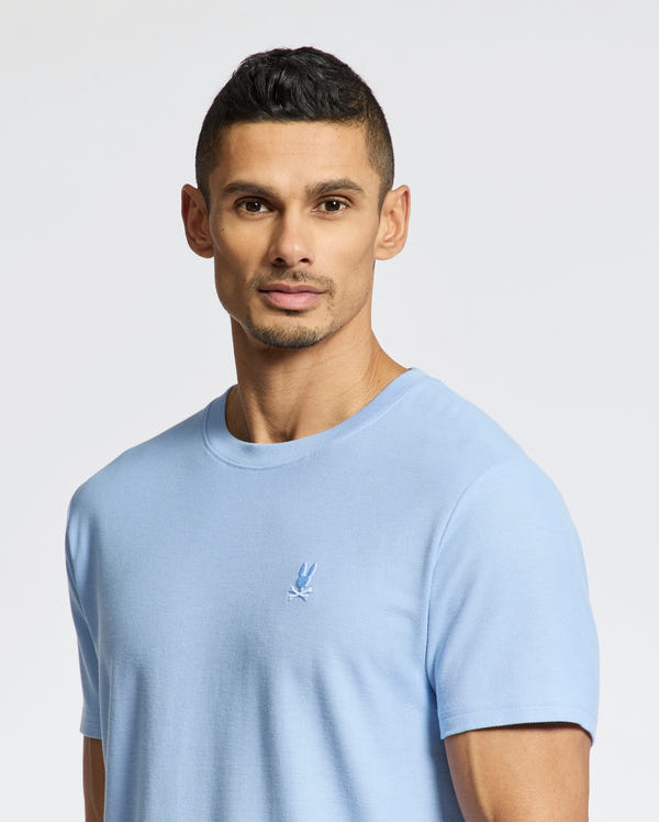 A person with short dark hair is wearing a light blue Psycho Bunny Men's Taylor Pique Tee (B6U579F200) featuring an embroidered chest Bunny. They stand against a plain background, looking at the camera.