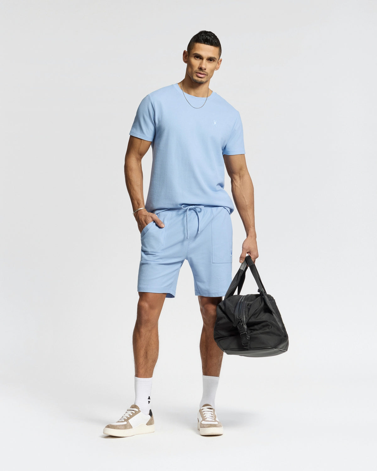 A man in a light blue Psycho Bunny MENS TAYLOR PIQUE TEE (B6U579F200) with matching shorts stands holding a black duffel bag, sporting short hair, white socks, and beige sneakers. An embroidered chest Bunny enhances the outfit's charm against a plain white background.
