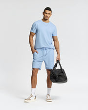 A man in a light blue Psycho Bunny MENS TAYLOR PIQUE TEE (B6U579F200) with matching shorts stands holding a black duffel bag, sporting short hair, white socks, and beige sneakers. An embroidered chest Bunny enhances the outfit's charm against a plain white background.