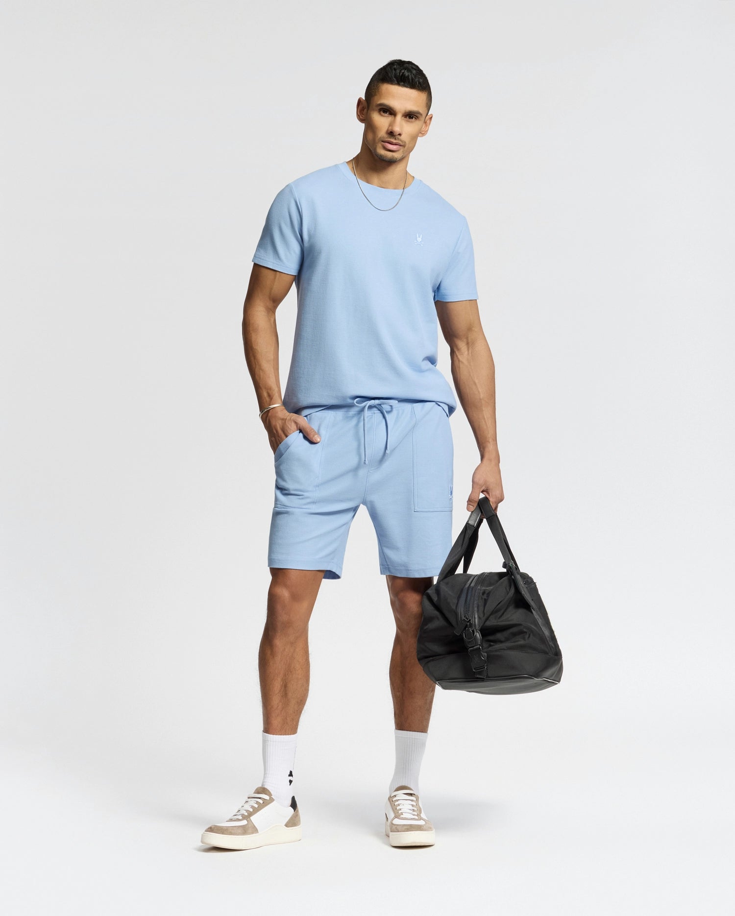 A man in a light blue Psycho Bunny MENS TAYLOR PIQUE TEE (B6U579F200) with matching shorts stands holding a black duffel bag, sporting short hair, white socks, and beige sneakers. An embroidered chest Bunny enhances the outfit's charm against a plain white background.