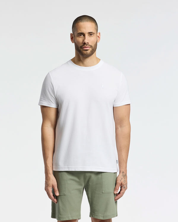 A short-haired, bearded individual stands against a white backdrop wearing a Psycho Bunny MENS TAYLOR PIQUE TEE in regular fit, crafted from lightweight Pima cotton, complemented by olive green shorts. Their relaxed stance is highlighted by the tee's embroidered chest bunny.