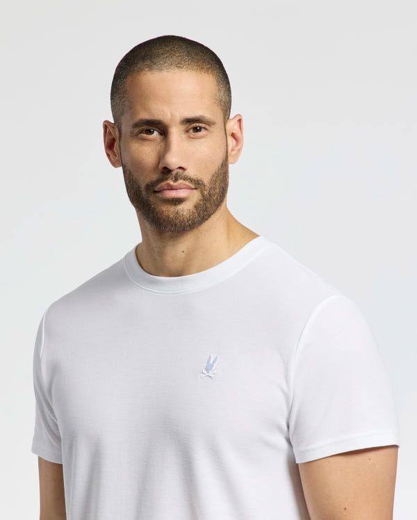 A man with a shaved head and beard wears the Psycho Bunny Men's Taylor Pique Tee (B6U579F200), made of lightweight Pima cotton for comfort. The plain white tee features a small embroidered bunny on the chest. He stands against a neutral gray background, gazing at the camera.