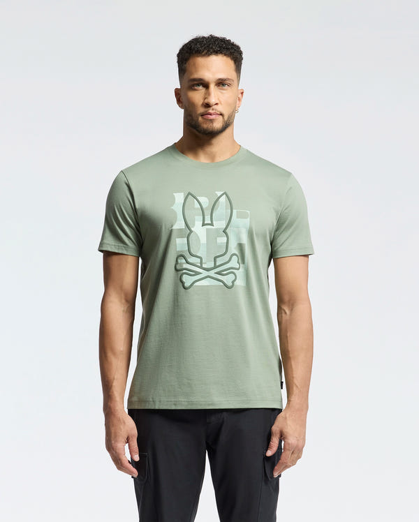 A man faces forward against a plain light background, wearing Psycho Bunny's MENS ZEALAND GRAPHIC TEE (B6U604F200) in light green with a large bunny head and crossbones graphic on the front, paired with black pants.