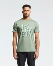 A man faces forward against a plain light background, wearing Psycho Bunny's MENS ZEALAND GRAPHIC TEE (B6U604F200) in light green with a large bunny head and crossbones graphic on the front, paired with black pants.
