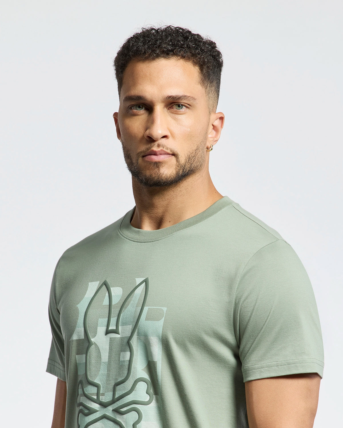 A man with short curly hair, a trimmed beard, and a gold hoop earring wears the Psycho Bunny MENS ZEALAND GRAPHIC TEE (B6U604F200) in sage green, featuring a bunny and crossbones graphic, standing against a plain light background.