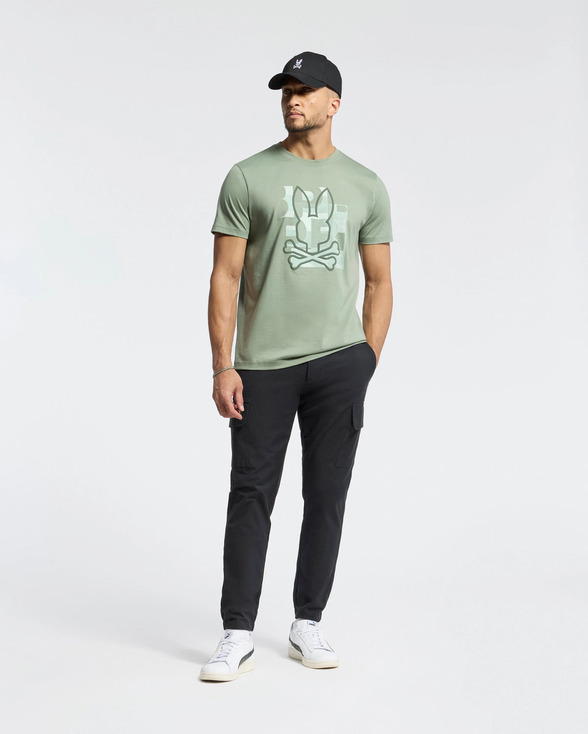 A man in a Psycho Bunny MENS ZEALAND GRAPHIC TEE (B6U604F200) featuring a large Bugs Bunny face graphic, black cap, black pants, and white sneakers stands against a plain white background.
